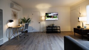 Ein modernes Wohnzimmer mit Flachbildfernseher an der Wand, einem Schreibtisch mit Stuhl, einer Couch und einem kleinen Beistelltisch mit Lampe.