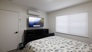 Ein Schlafzimmer mit Bett, Fernseher an der Wand, Fenster mit Jalousien und einer Tür.