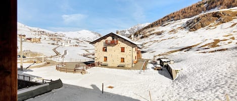 A snow-covered mountain village with a wooden building featuring a balcony, surrounded by snow-covered terrain and a clear sky.