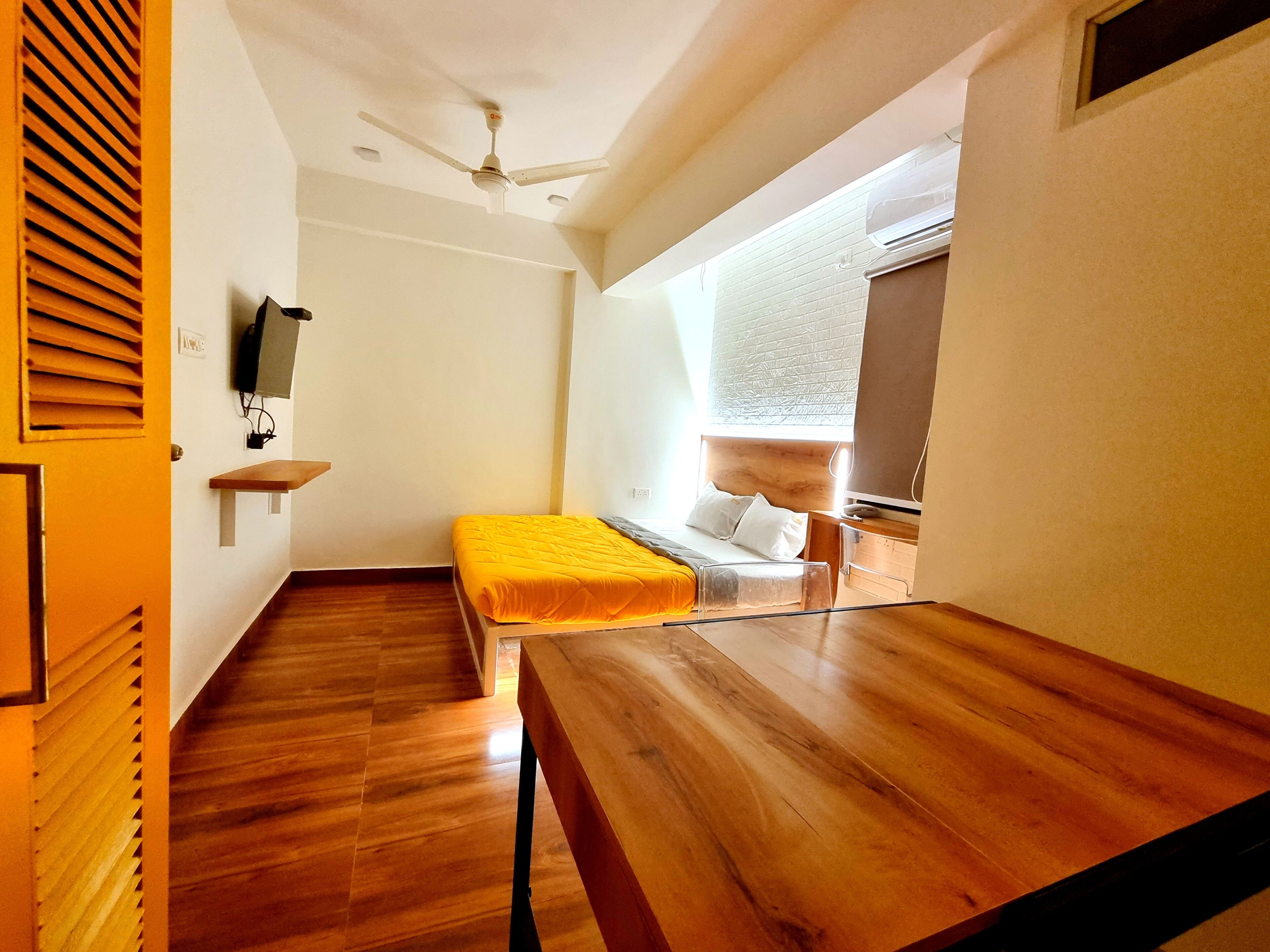 A room with a bed, a wooden table, a ceiling fan, a window with blinds, and a wall-mounted TV.