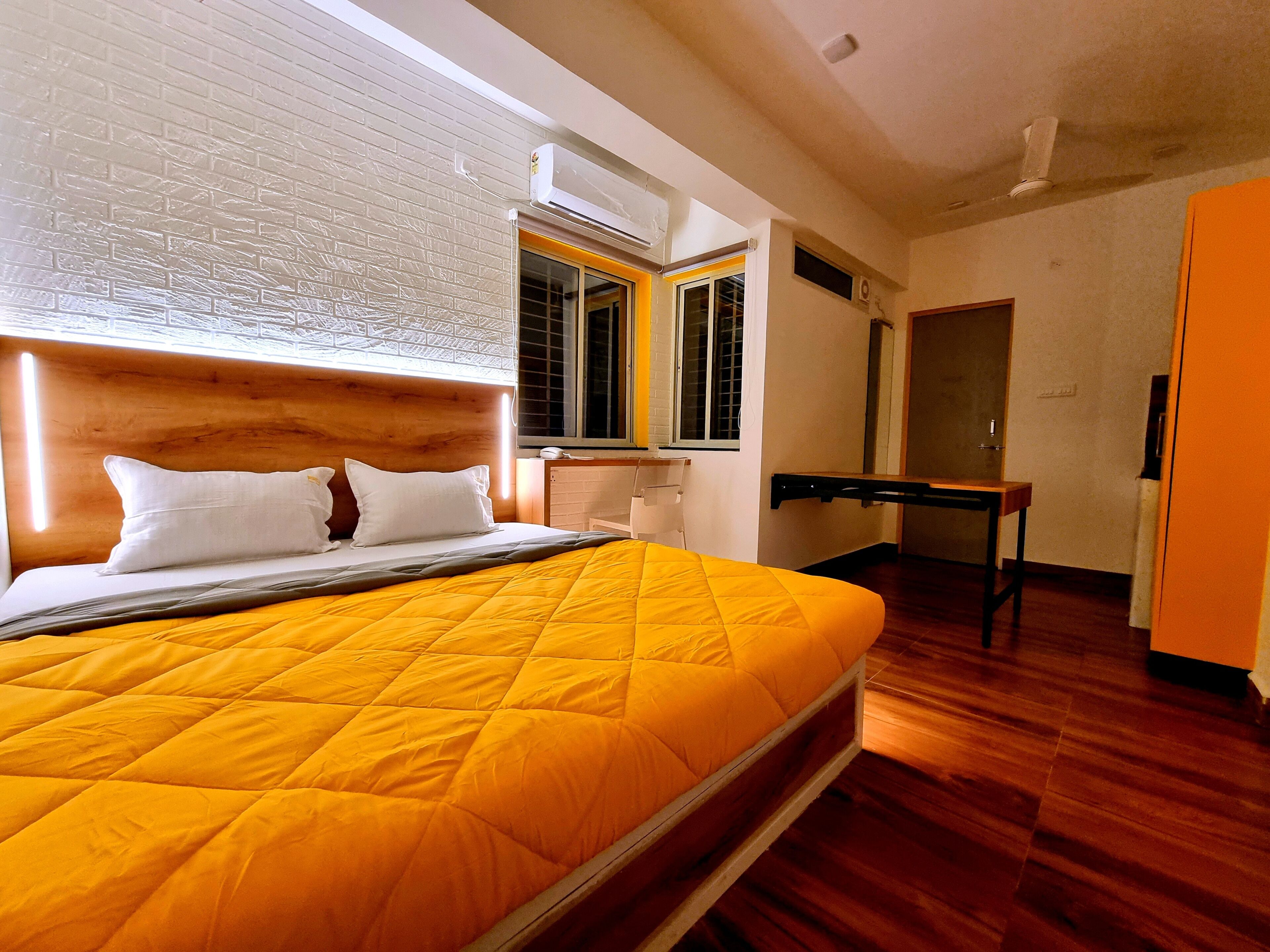 A hotel room with a large bed, a wooden headboard, an air conditioning unit, a window with blinds, a bedside table, and a door.