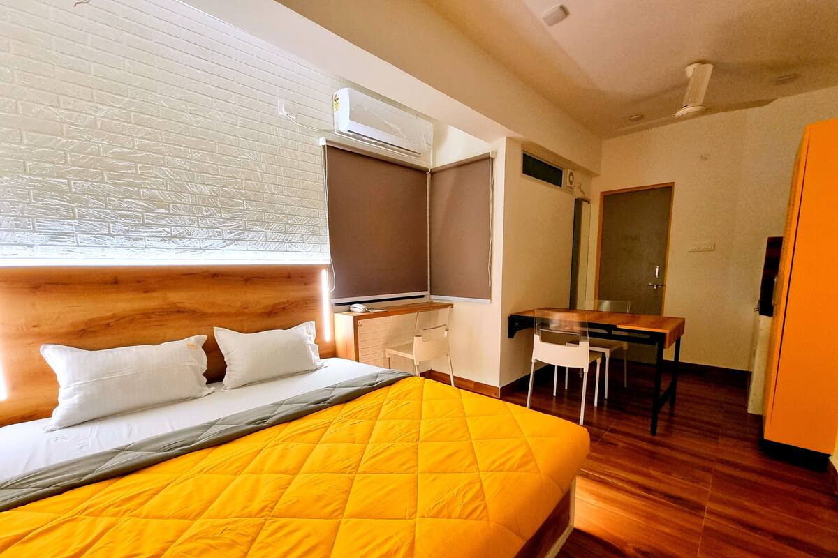 A hotel room with a bed, a desk, a chair, an air conditioning unit, and a window with blinds.