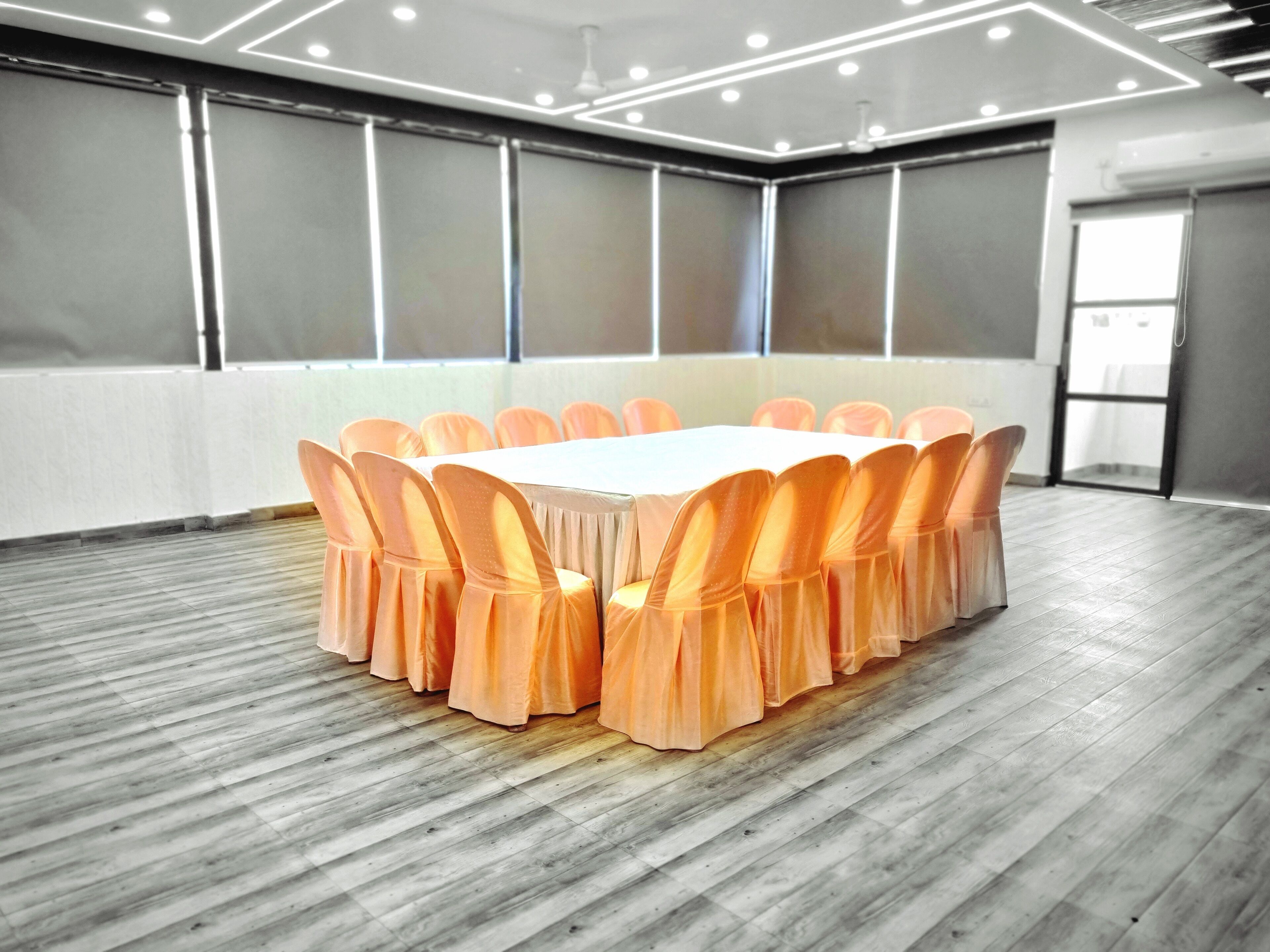 A conference room with a long table surrounded by chairs, a wooden floor, and a large window with blinds.