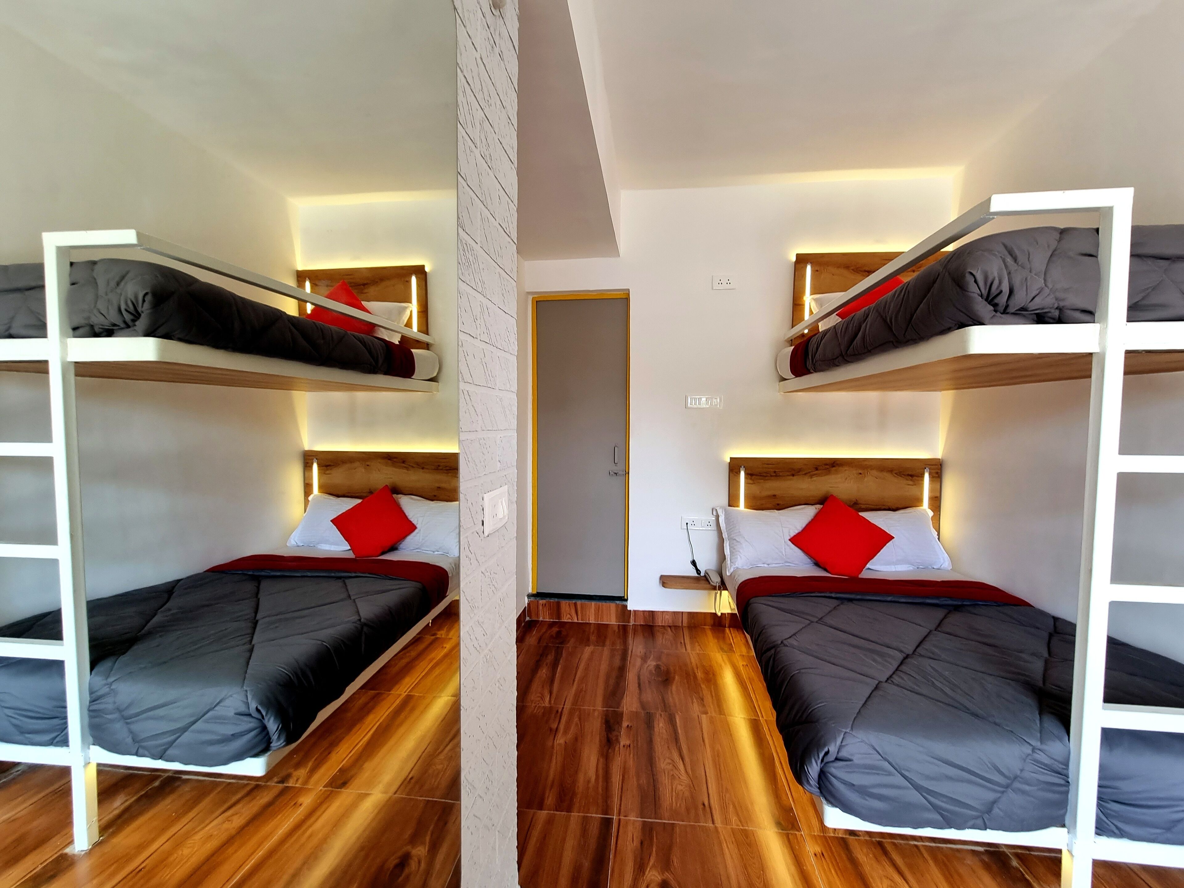 A room with two bunk beds, each with a wooden headboard and a red pillow. The room has wooden flooring and white walls with a textured pattern.