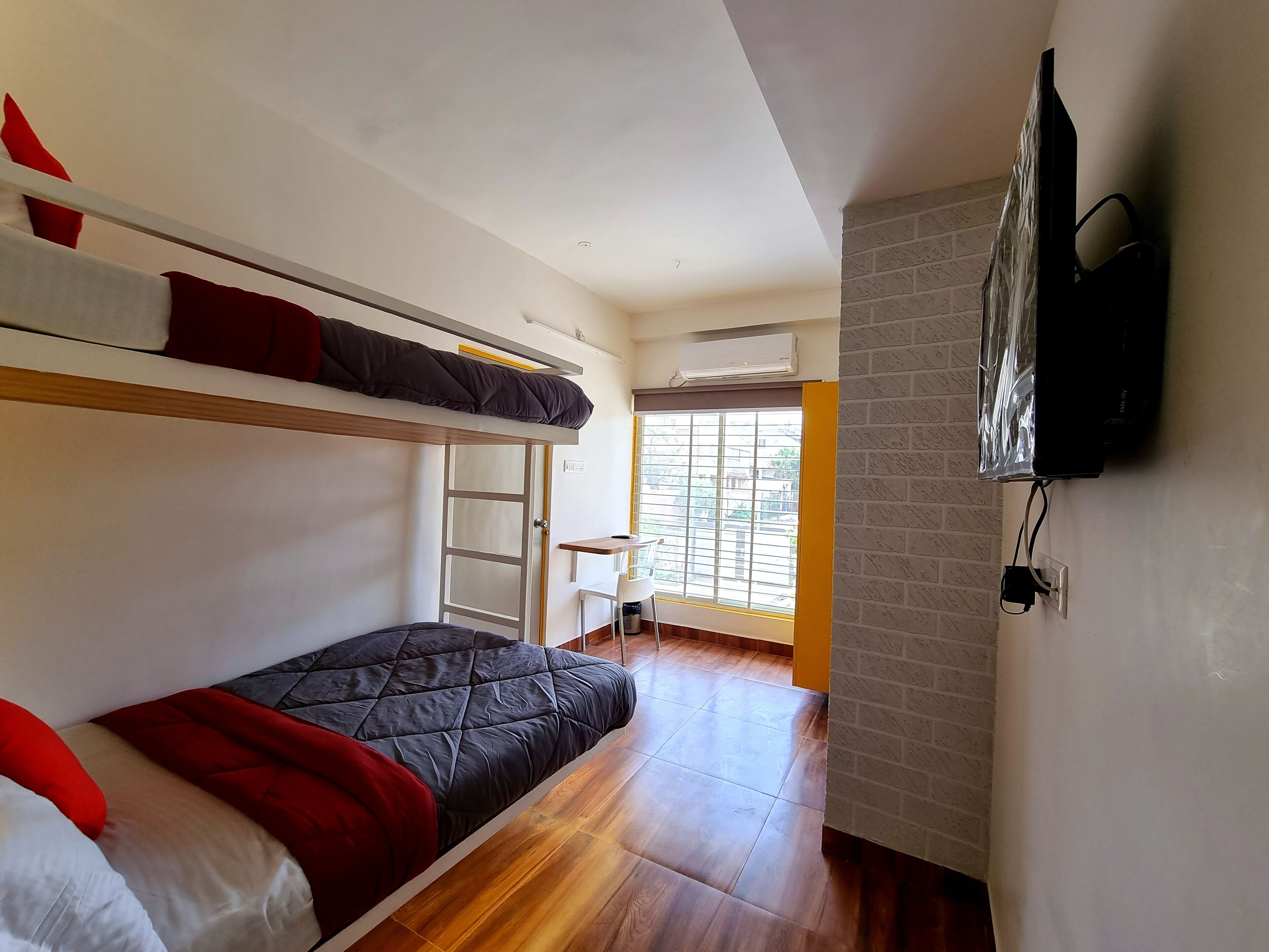 A dormitory room with bunk beds, a desk, a chair, and a television mounted on the wall.