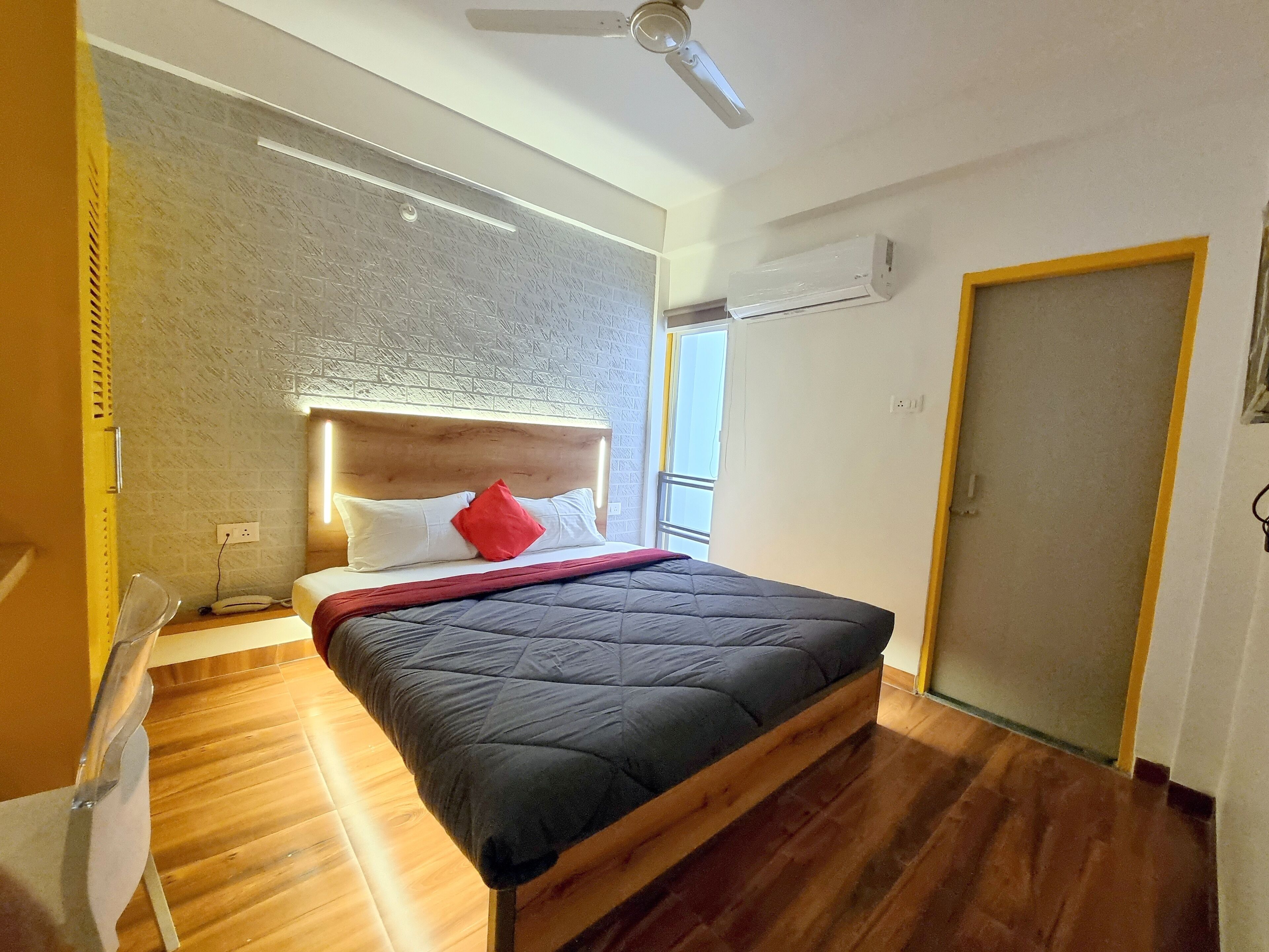 A modern bedroom with a wooden headboard, a large bed with a dark quilt, a red pillow, and an air conditioning unit on the wall.