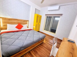 Ein Hotelzimmer mit einem hölzernen Kopfteil, einer grauen gesteppten Bettdecke, einem roten Kopfkissen und einem gelben Kleiderschrank. Ein Fenster mit Jalousien ist ebenfalls zu sehen.