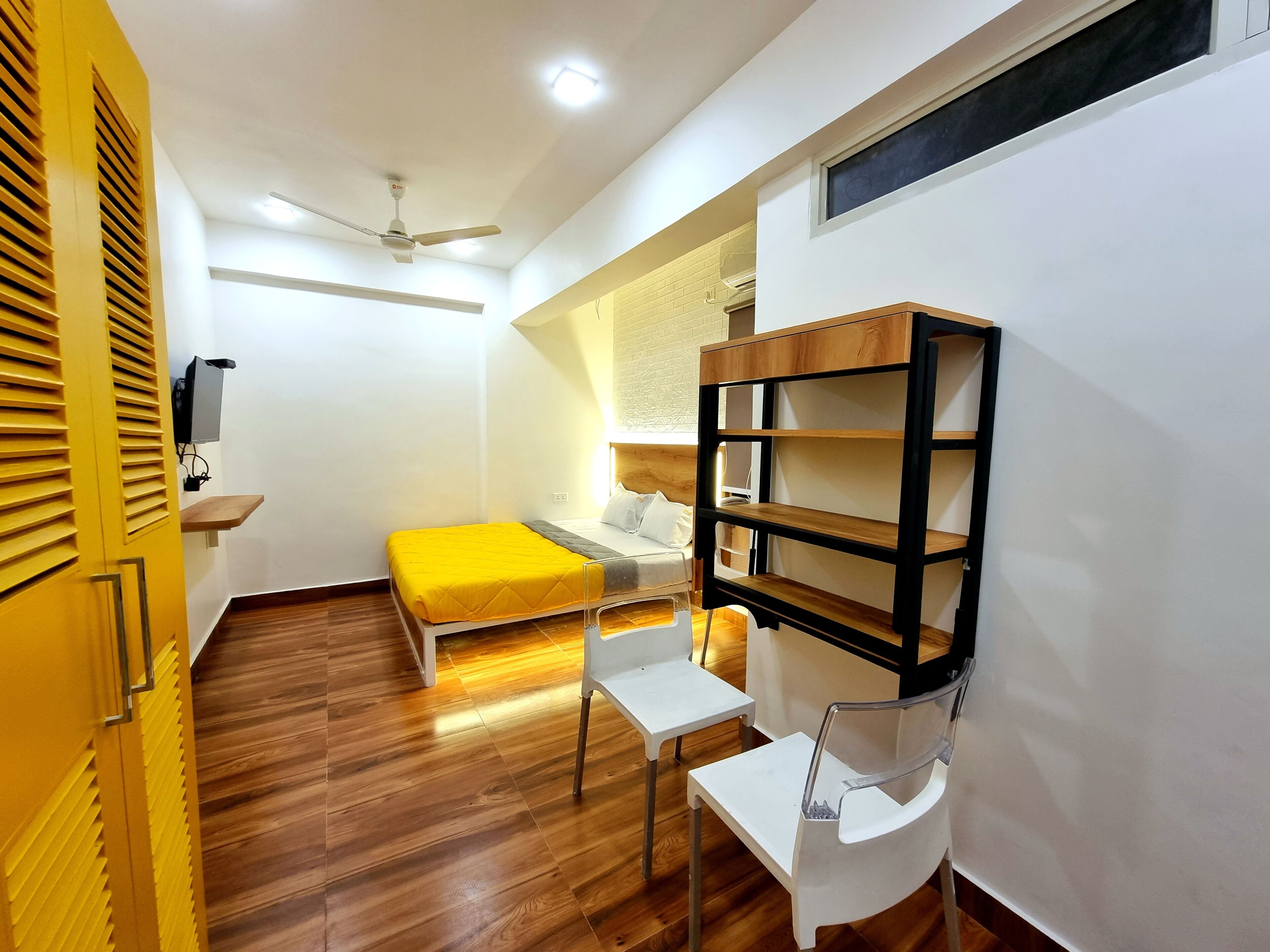 A compact room with a bed, a desk, a chair, a ceiling fan, and a shelf unit.