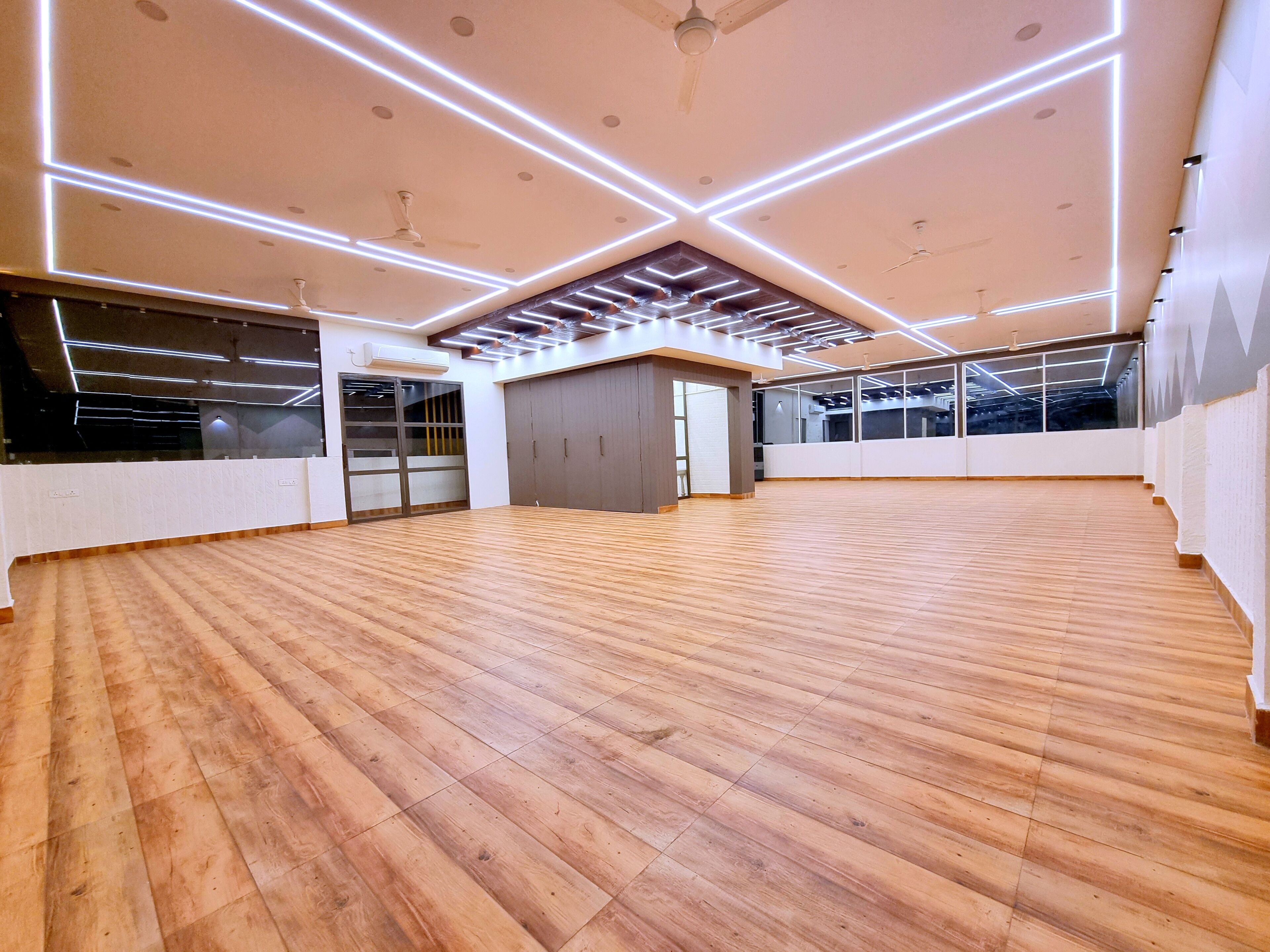 A spacious room with wooden flooring, large windows, and a ceiling with linear lighting.