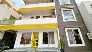 A multi-story residential building with a yellow and white facade, a visible entrance with a yellow door, and a small garden area with potted plants. - Stay inn nest (Bhopal)