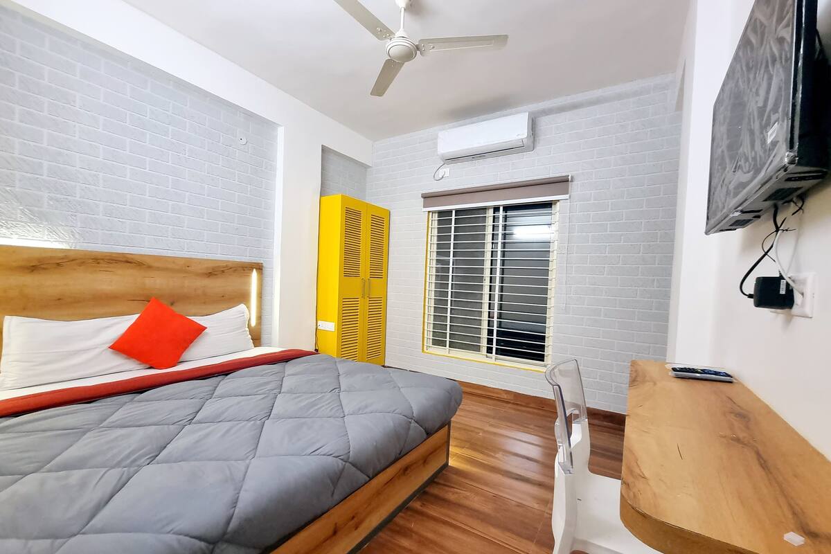 A modern bedroom with a wooden bed, a grey quilted mattress, a red pillow, a white bedside table, a wooden chair, a yellow wardrobe, a window with blinds, a ceiling fan, and an air conditioning unit.