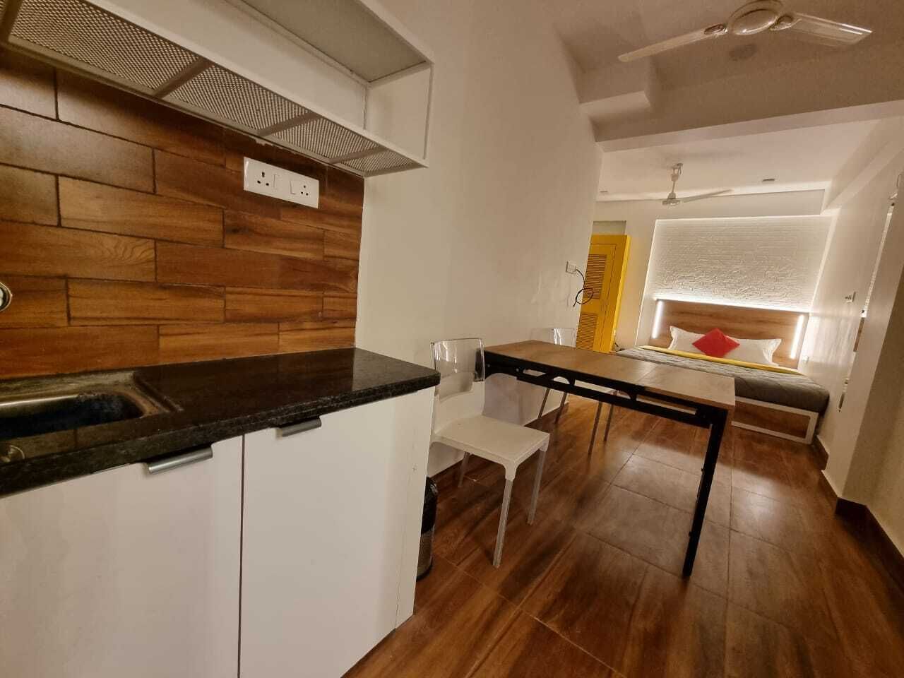 Uma cozinha compacta com painel de parede de madeira, armários brancos e bancada preta. Há uma área de refeições com mesa e cadeiras, e uma cama com cabeceira ao fundo.