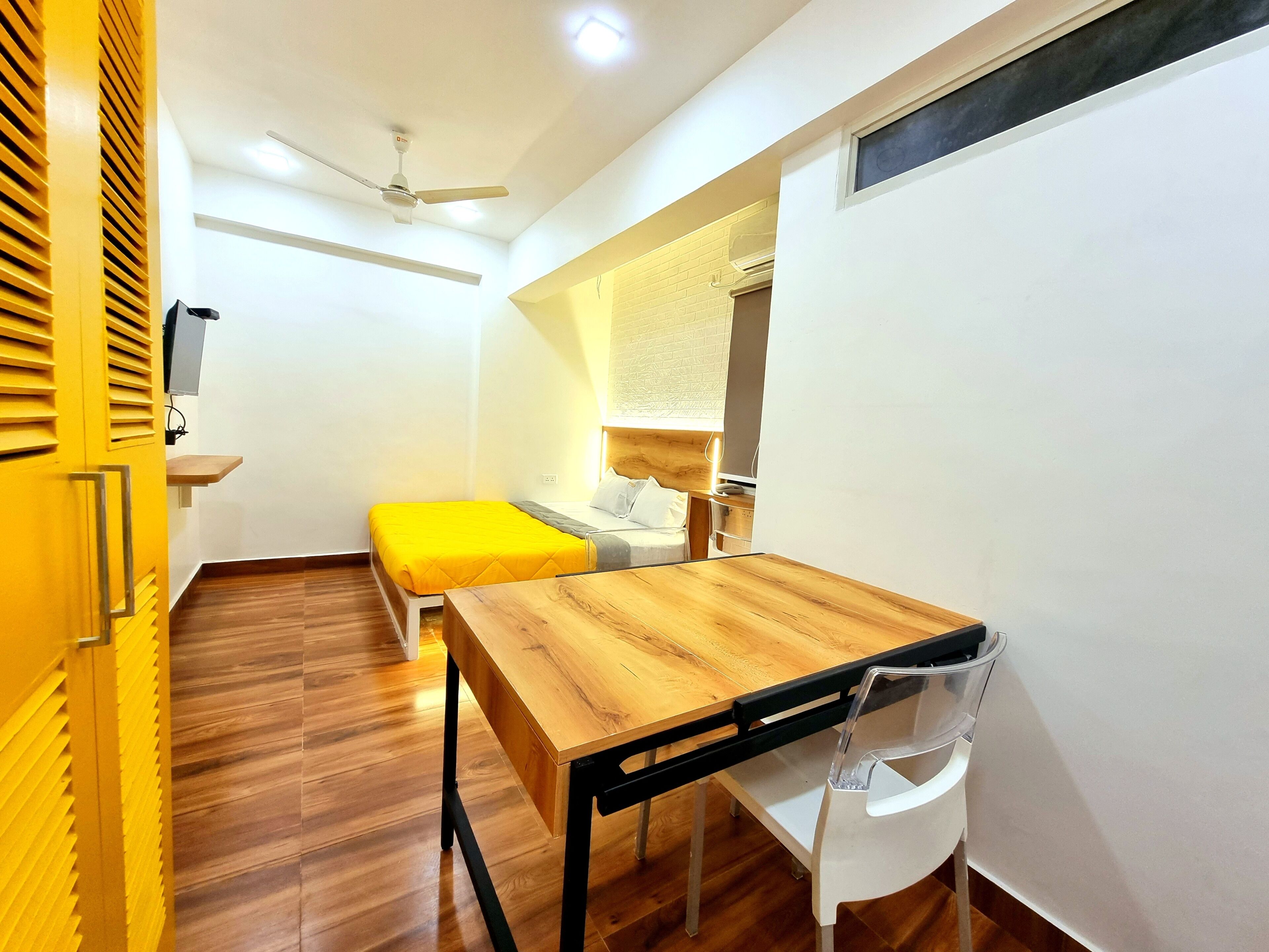 A compact room with a wooden desk, a single bed with a yellow bedspread, a ceiling fan, and a window with blinds.