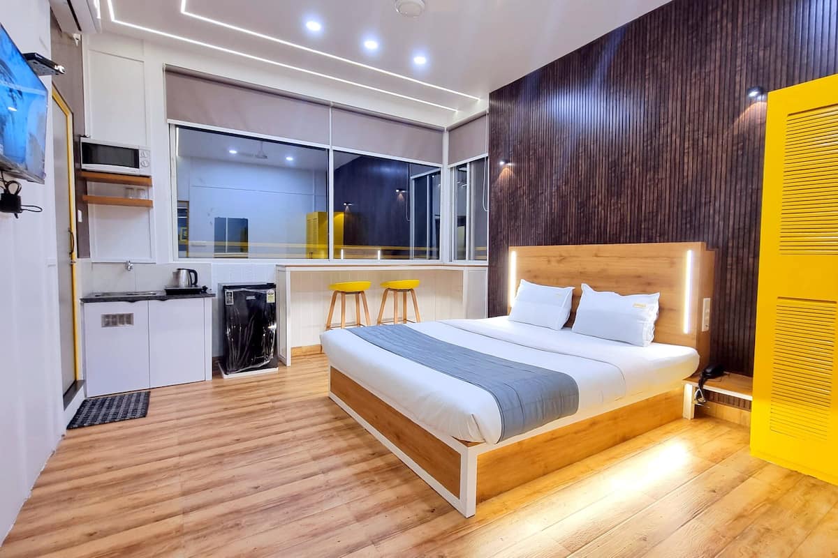 A modern hotel room with a wooden floor, a bed with white bedding, a small kitchenette, and a large window.