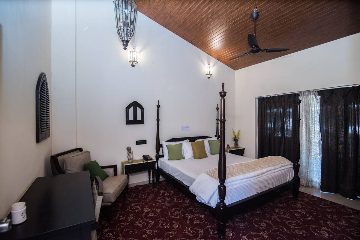 A four-poster bed with a wooden headboard, a nightstand with a lamp, a chair, and a desk with a telephone.