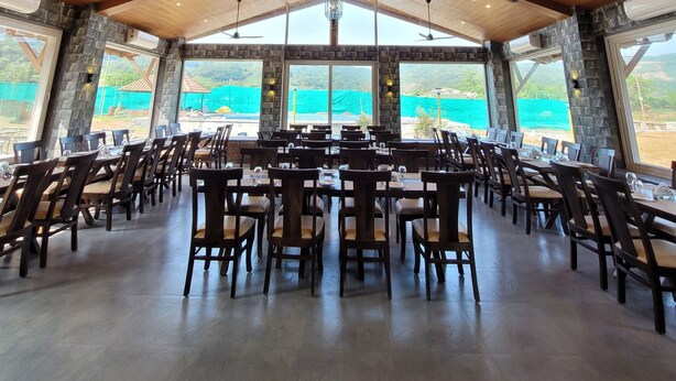 A spacious dining area with wooden ceiling beams, large windows offering a view of the outdoors, and multiple ceiling fans. - Mastiff Grand La Villae Resort (Mawal)
