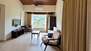 A spacious living area with a wooden ceiling, a ceiling fan, a TV mounted on the wall, a sofa, a coffee table, and a view of the outdoors through large windows. - Mastiff Grand La Villae Resort (Mawal)
