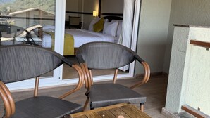 A balcony with wicker chairs and a wooden table, a bedroom with a bed and curtains, and a mountain view through a glass door. - Mastiff Grand La Villae Resort (Mawal)
