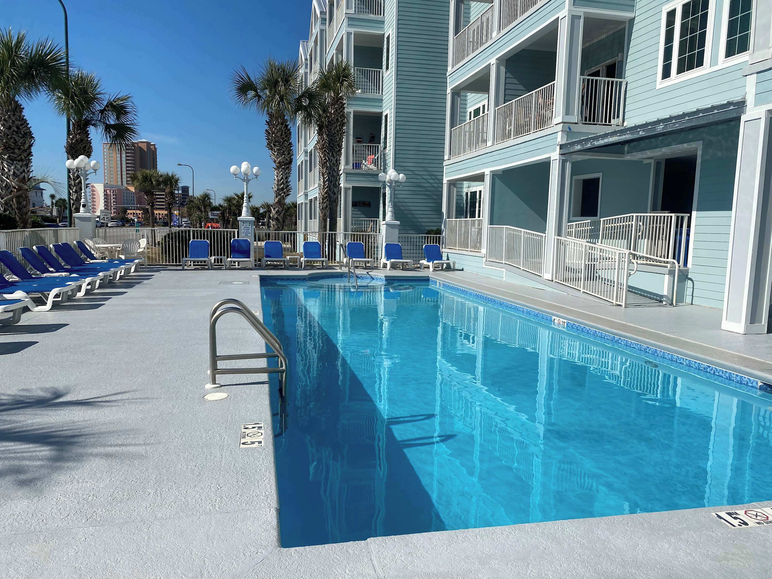 Uma piscina com água azul, rodeada por espreguiçadeiras e um edifício com varandas.