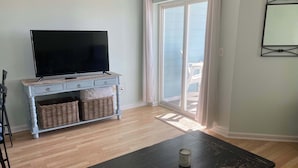 A living room with a flat-screen TV on a light-colored console, wicker baskets, a dark wooden coffee table, and a sliding glass door leading to a balcony. - Grand Caribbean 205 - Condo has beautiful views of the Gulf and great rates! (Orange Beach)