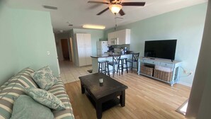 A living room with a sofa, a coffee table, and a TV stand with a flat-screen TV. There is a kitchen area with cabinets and a ceiling fan. - Grand Caribbean 205 - Condo has beautiful views of the Gulf and great rates! (Orange Beach)