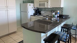 A kitchen with white cabinets, a refrigerator, a stove, a microwave, and a coffee maker. There are two bar stools and a sink. - Grand Caribbean 205 - Condo has beautiful views of the Gulf and great rates! (Orange Beach)