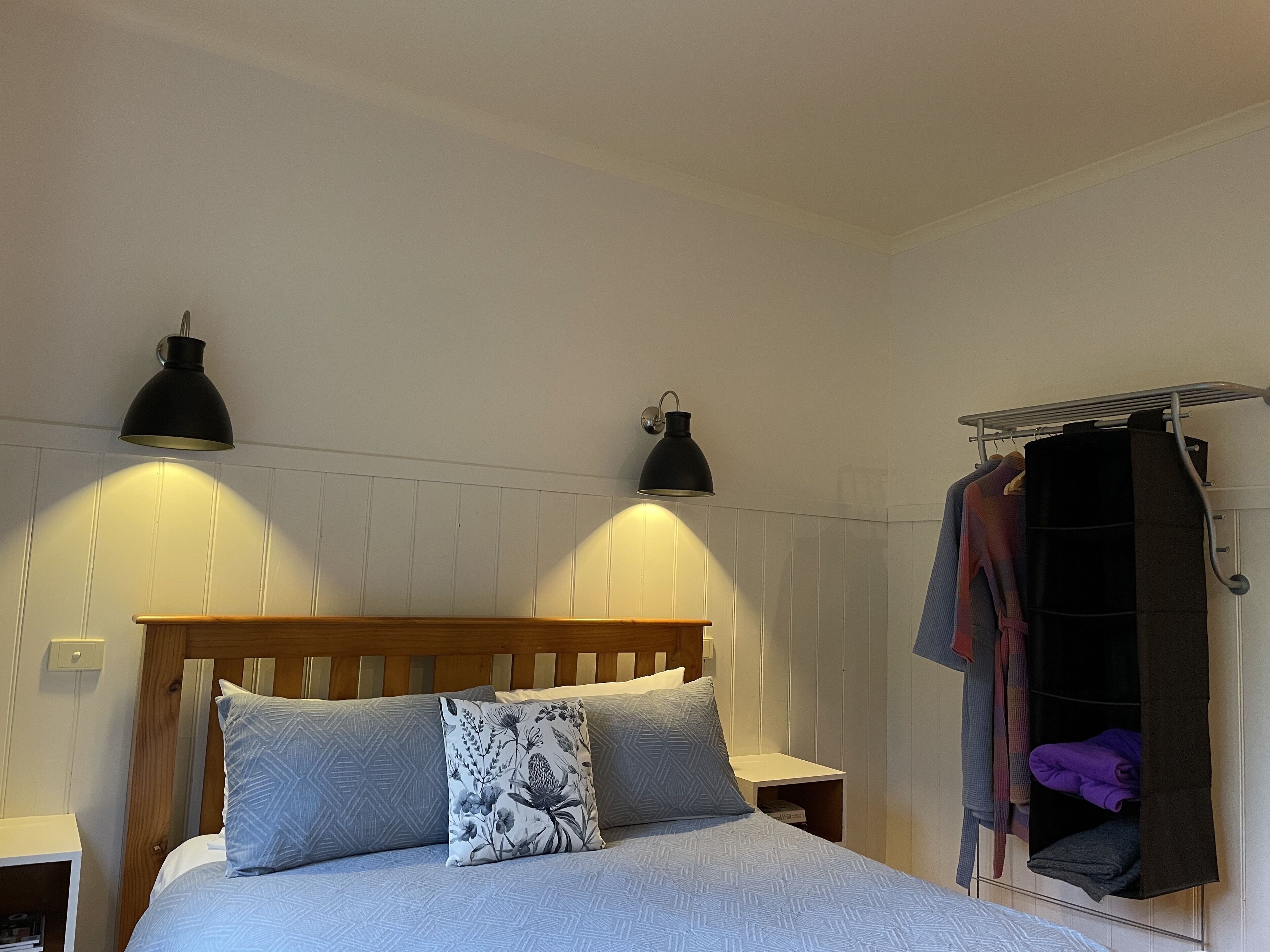 Un dormitorio con cabecera de madera, dos lámparas de noche, una cama con ropa de cama estampada, una mesita de noche y un perchero con ropa doblada.
