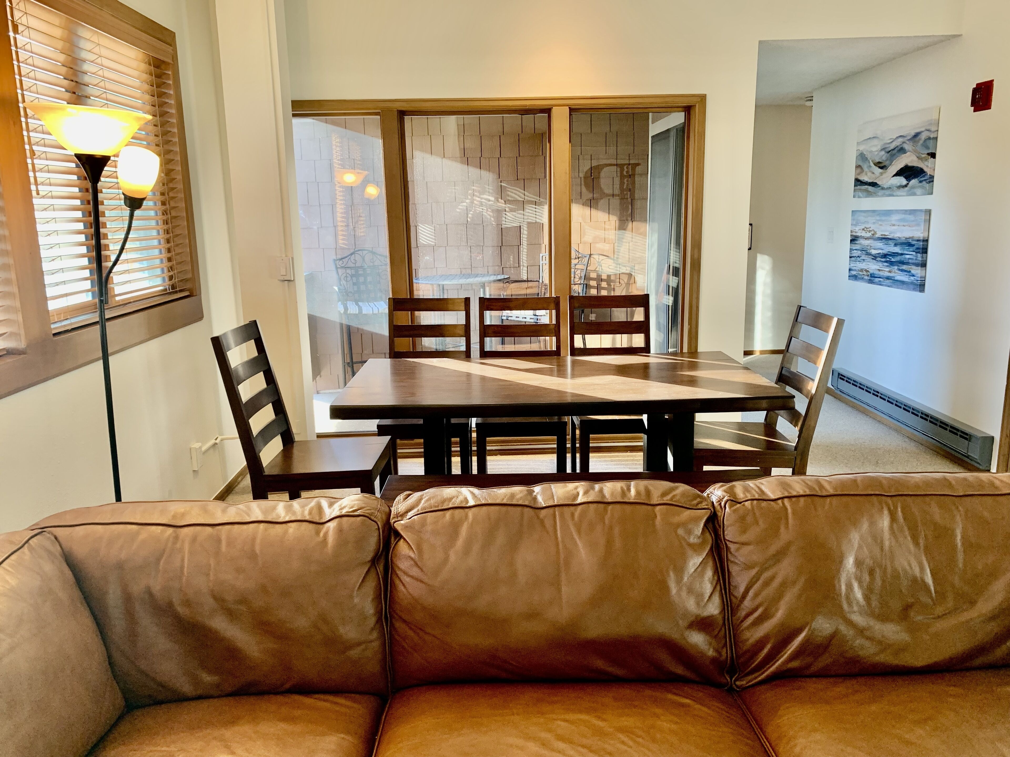 A dining area with a wooden table and chairs, a leather couch, and a view through a glass door.