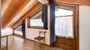Una habitación con techo inclinado, vigas de madera, una silla de madera, una cama y una ventana con cortinas.