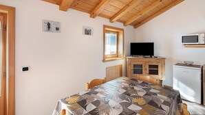Ein Zimmer mit Holzboden, einem Fernseher auf einem Schrank, einem Kühlschrank und einem Tisch mit gemustertem Tischläufer.