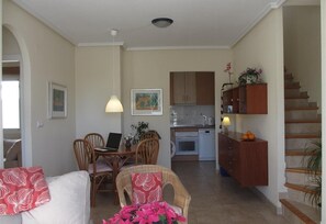 A living room with a dining table, chairs, a sofa, a kitchen in the background, and a staircase.