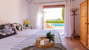 Quarto com cama, mesa de cabeceira com abajur, bandeja com xícaras e vista para piscina e área de estar externa.