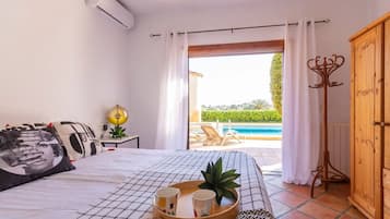 Une chambre avec un lit, une table de chevet avec une lampe, un plateau avec des tasses, et une vue sur une piscine et un espace de détente extérieur.