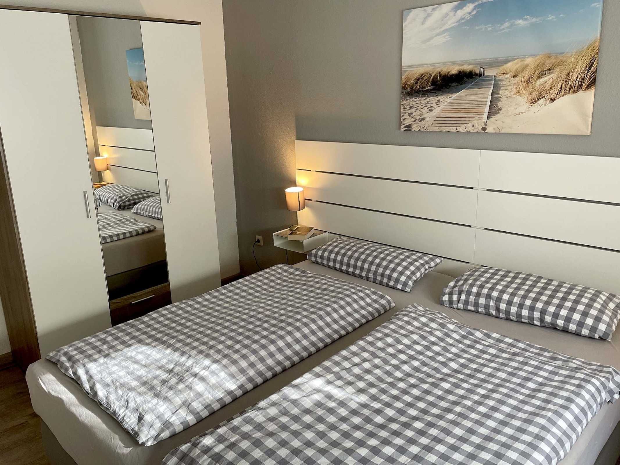 Ein Hotelzimmer mit Bett, Nachttisch, Lampe, Spiegel und Wandbild mit Strandmotiv.