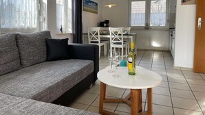Una sala de estar moderna con un sofá gris, una mesa de comedor blanca con sillas y una cocina al fondo.