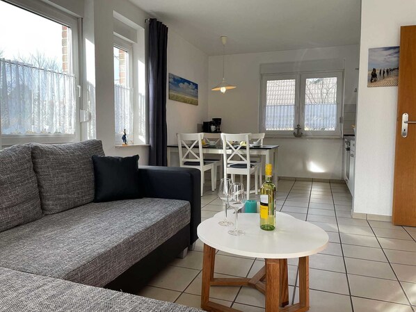 A modern living room with a grey sofa, a white dining table with chairs, and a kitchen area in the background.