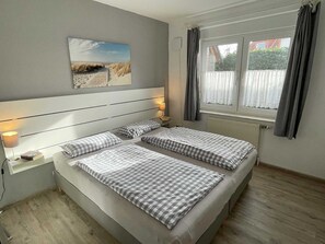 A bedroom with two single beds, a nightstand with a lamp, a window with curtains, and a beach-themed wall art.