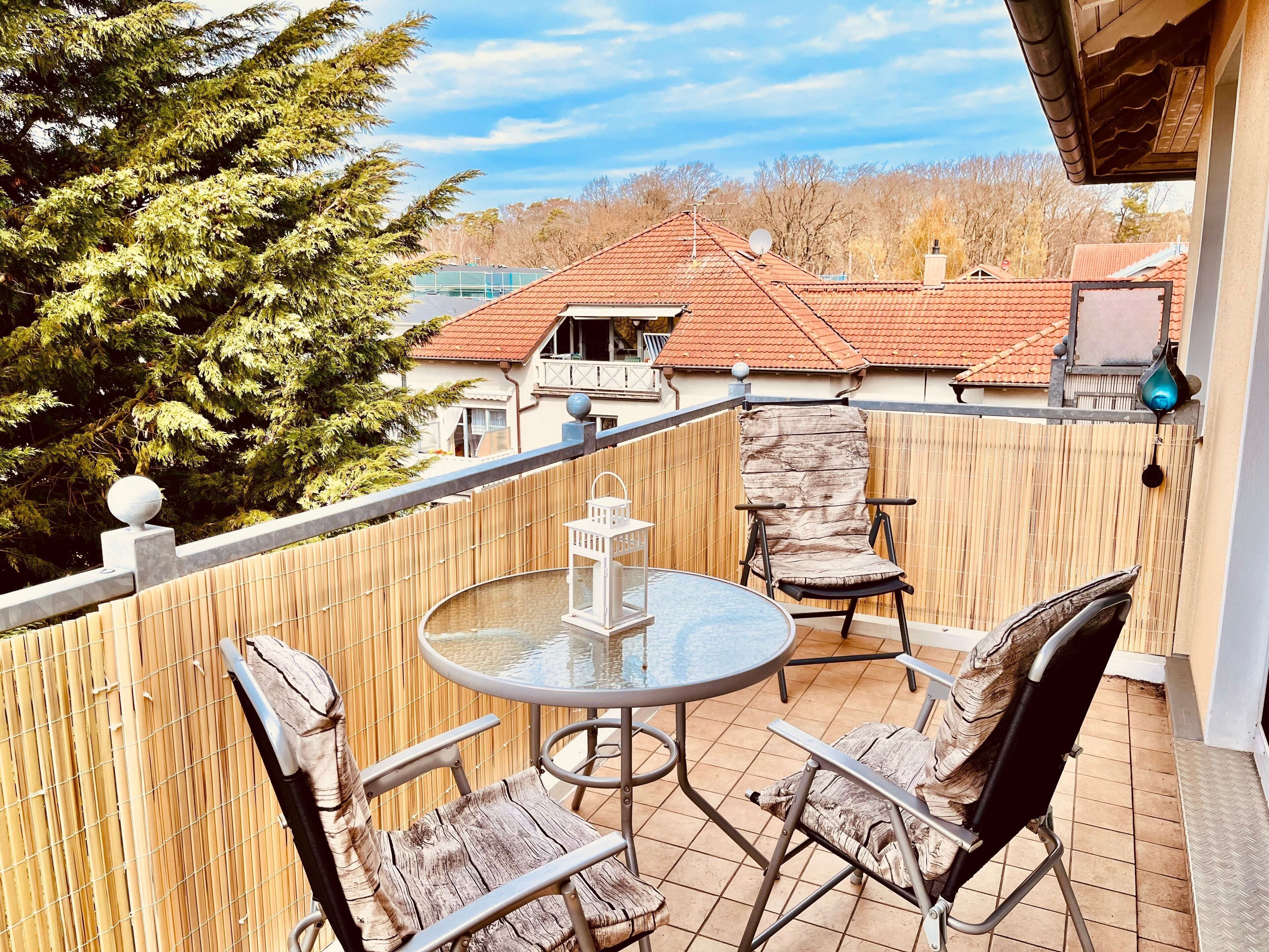 A balcony with a round table and two chairs, a wooden fence, and a view of houses and trees.