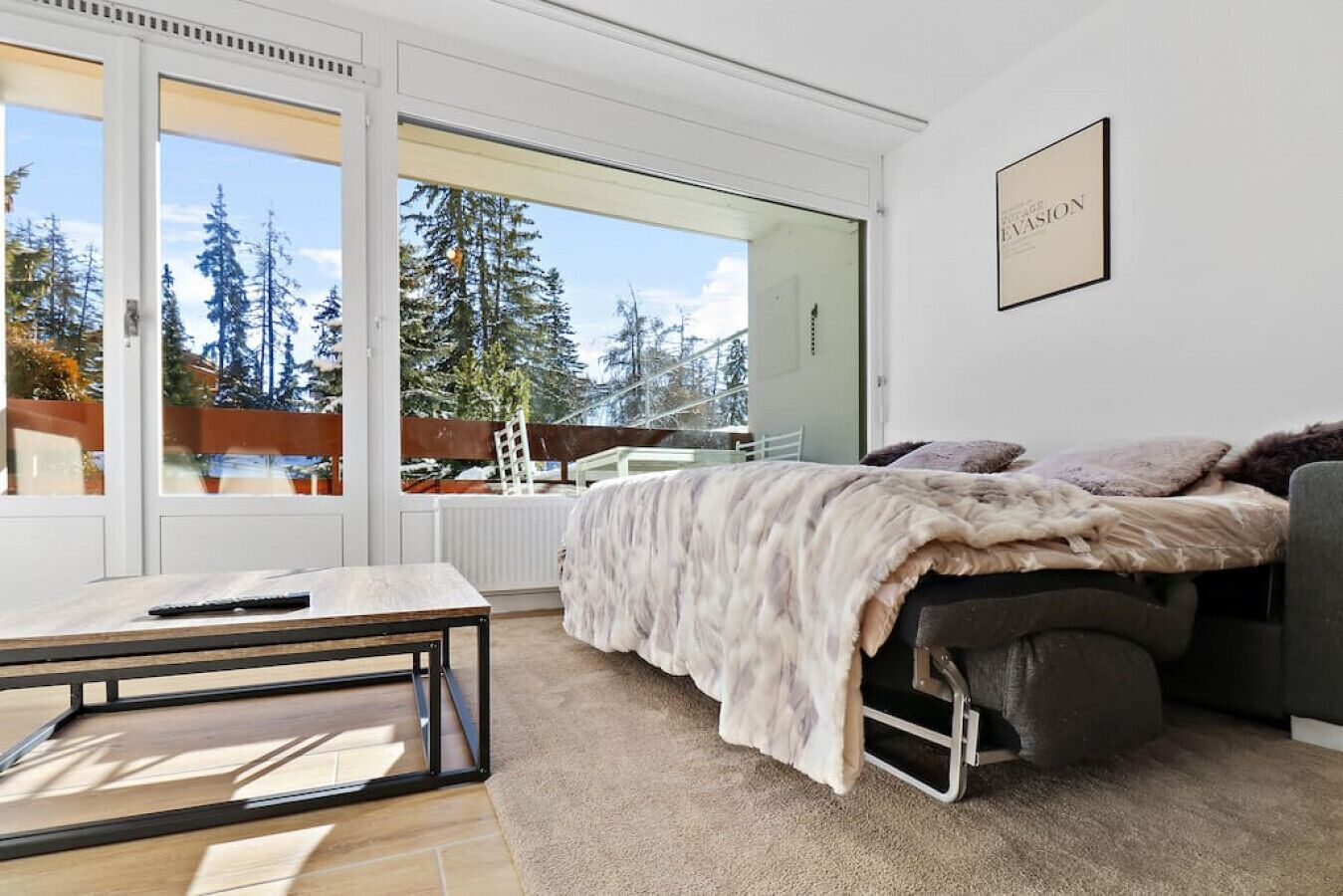 Un dormitorio moderno con cama, una mesita y vistas a árboles y una terraza de madera.