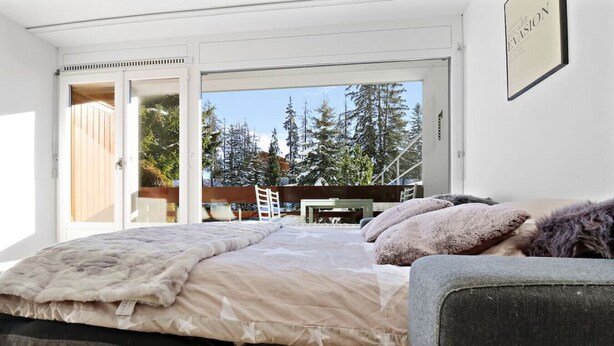A bedroom with a large bed, a view of trees and a wooden deck outside. - Cosy, renovated apartment in the heart of the resort (Crans-Montana)