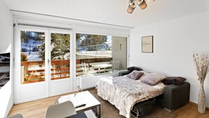 A modern bedroom with a large sliding glass door leading to a balcony, a sofa bed with a fur throw, a small table, and a chair. - Cosy, renovated apartment in the heart of the resort (Crans-Montana)