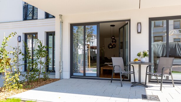 Un patio exterior moderno con una mesa y sillas, una puerta corrediza de vidrio que da a un espacio interior y una pequeña área de jardín con árboles jóvenes.