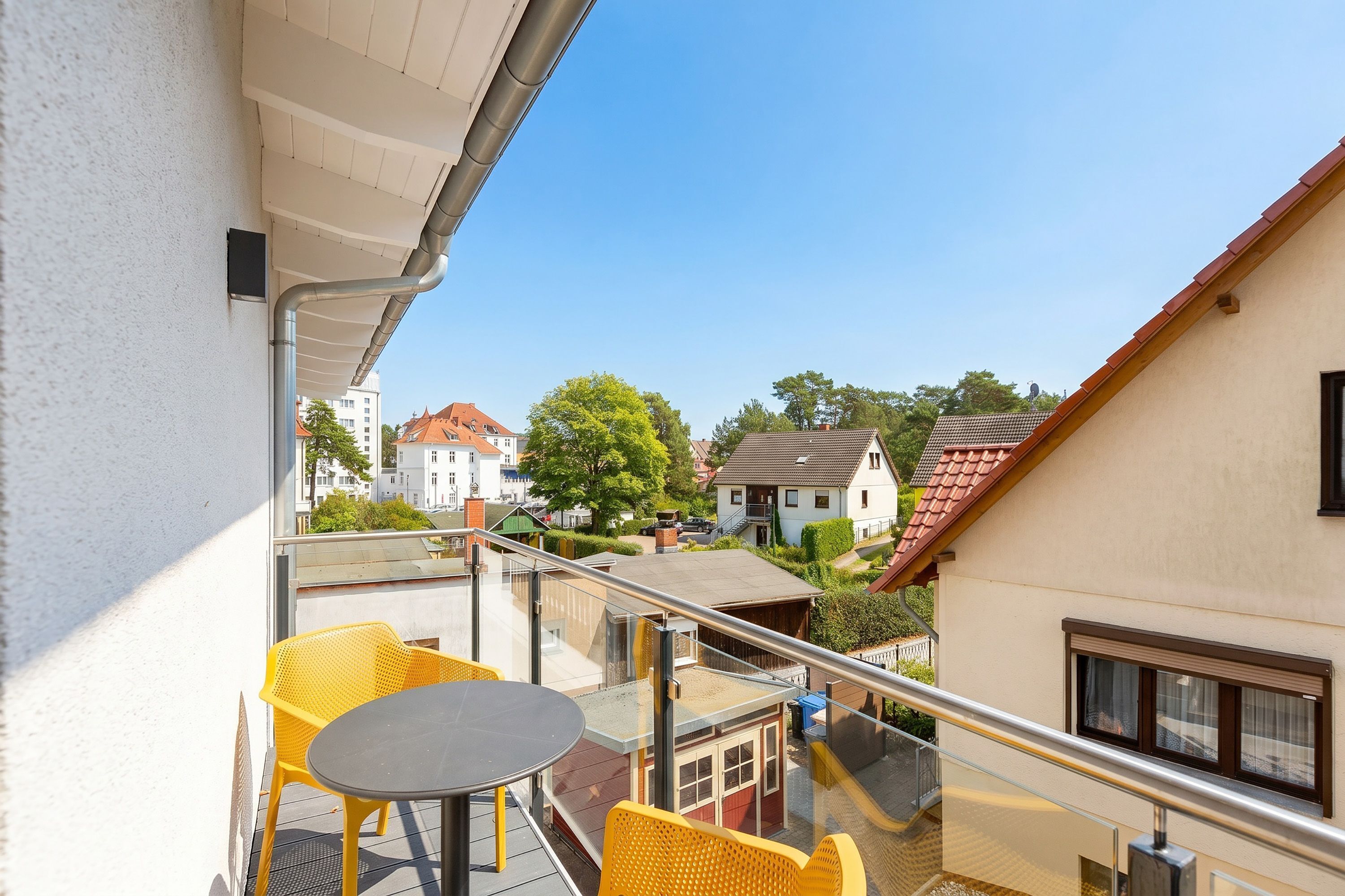 A balcony with yellow chairs and a table, overlooking a residential area with houses and trees.
