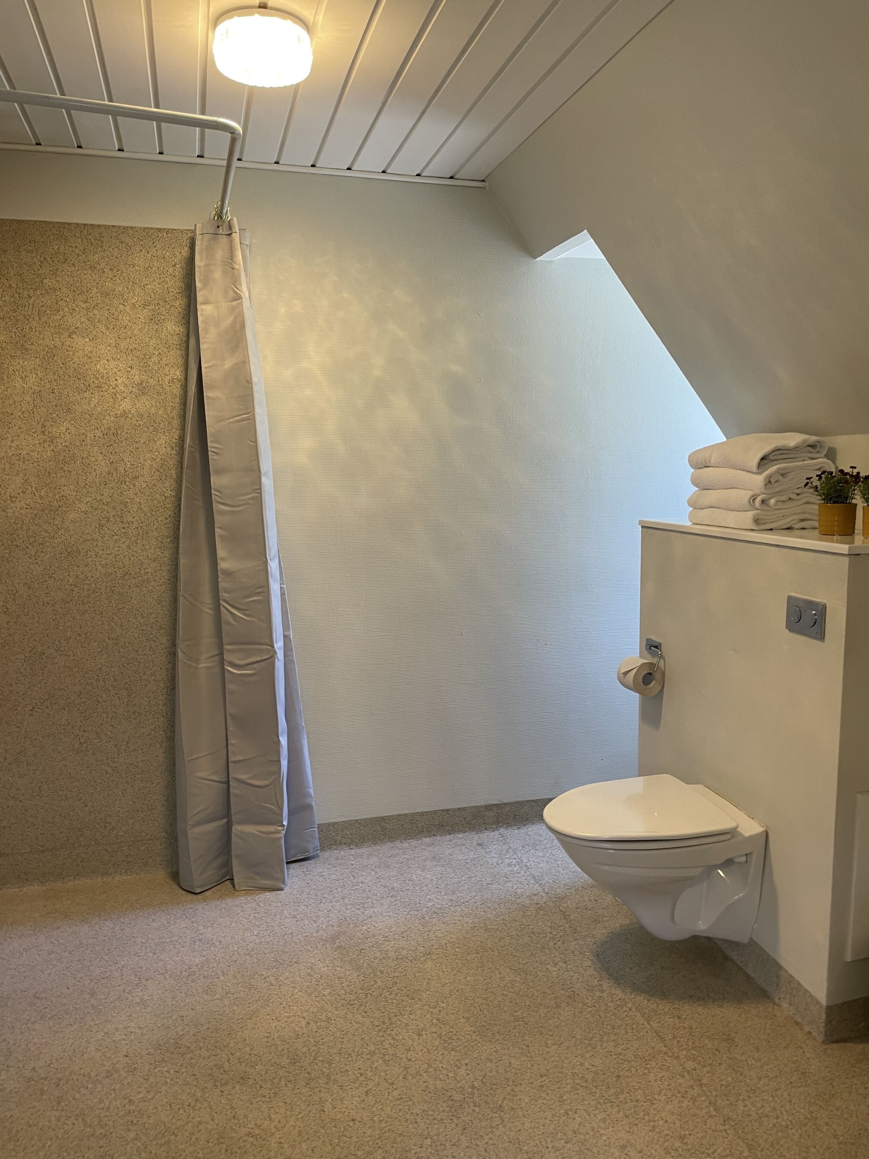 A small, compact bathroom with a toilet, a shower curtain, and a stack of towels on a shelf.