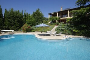 Ein Schwimmbecken mit Liegestühlen und Sonnenschirm, umgeben von üppiger Vegetation und einem Gebäude mit Balkon.