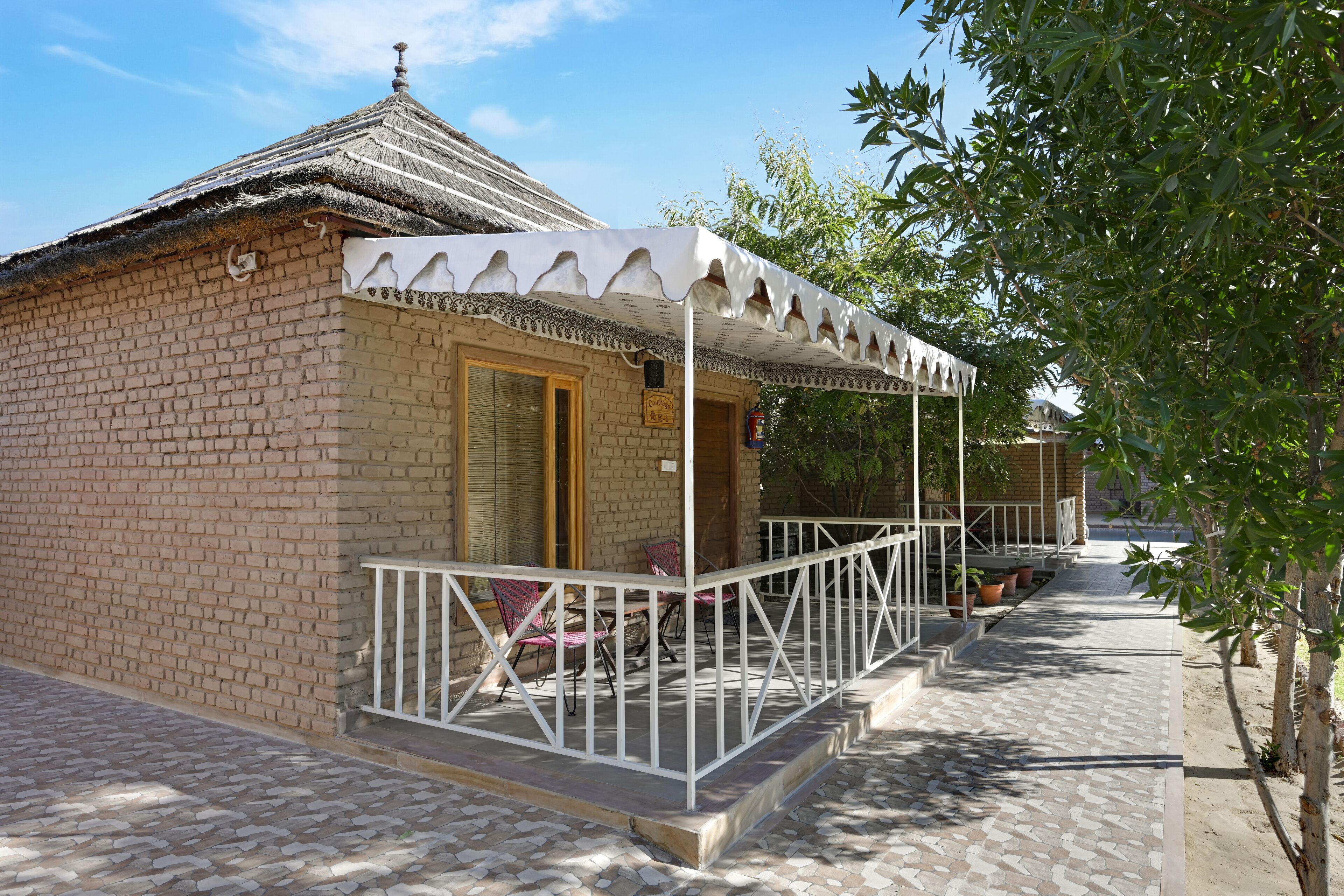 A small brick building with a thatched roof, a covered porch with white railings, and a paved walkway.