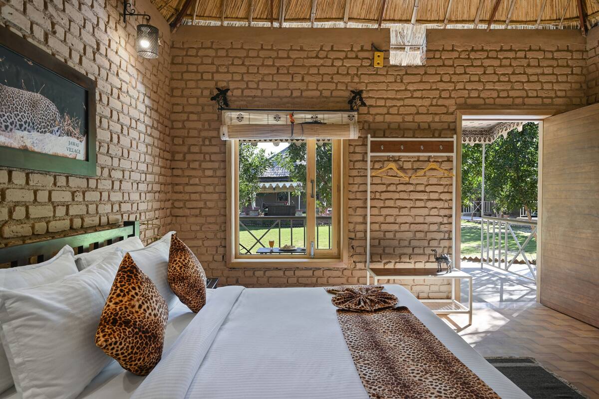 A bedroom with a brick wall, a bed with white linens, leopard print pillows, and a window with a view of the outdoors.