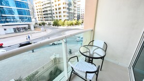 Un balcón con barandilla de vidrio que da a una calle de la ciudad, una mesa con dos sillas y vistas a edificios modernos.