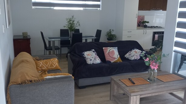 Ein modernes Wohnzimmer mit Sofa, Sessel und Couchtisch, ein Essbereich mit Stühlen und Tisch sowie eine Küche mit Schränken und Geräten.