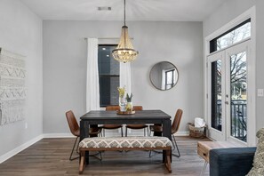 A dining area with a dark wooden table, six chairs, a bench, a hanging chandelier, a round mirror, and a window with a view.